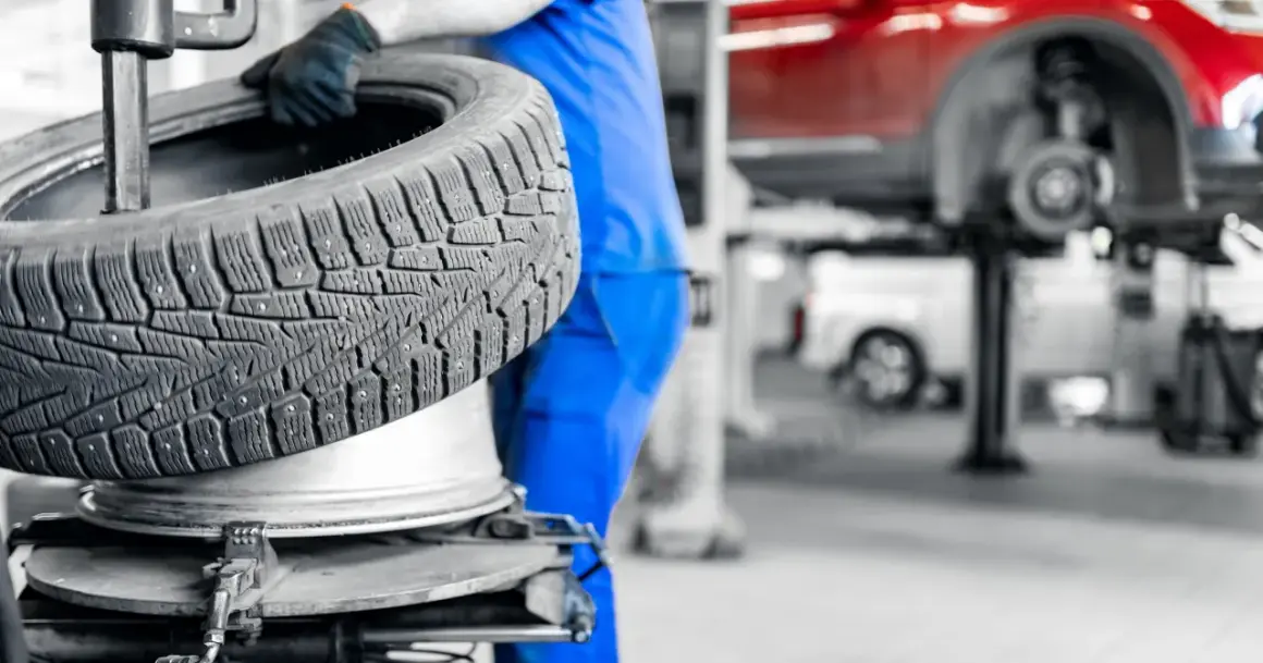 Mechanik w niebieskim kombinezonie trzyma oponę zimową. W tle widać podnośnik samochodowy i czerwony samochód. Sprawdź aktualne ceny wymiany opon.