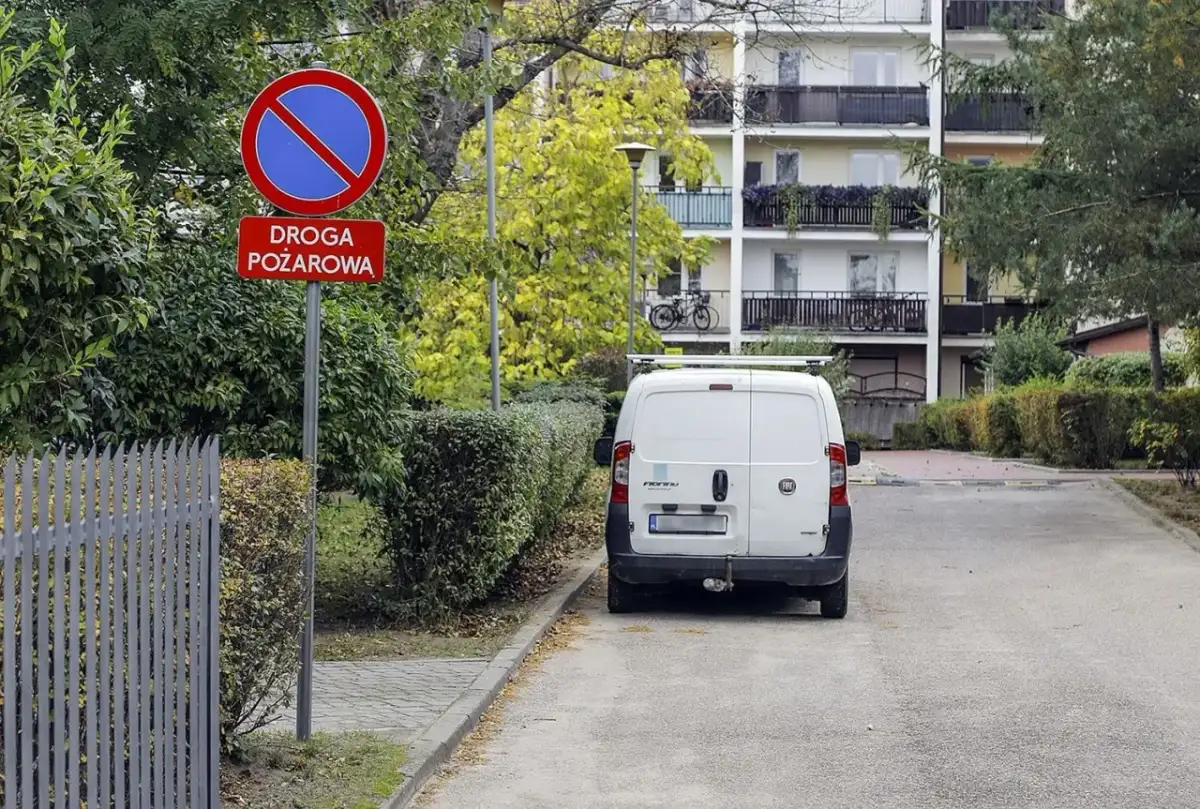 Znak zakazu postoju na drodze pożarowej. Biały bus stoi na poboczu, gdzie nie można parkować.