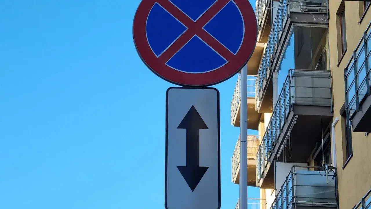 Znak zakazu zatrzymywania z tabliczką wskazującą dopuszczony ruch dwukierunkowy. Budynek z balkonami w tle.