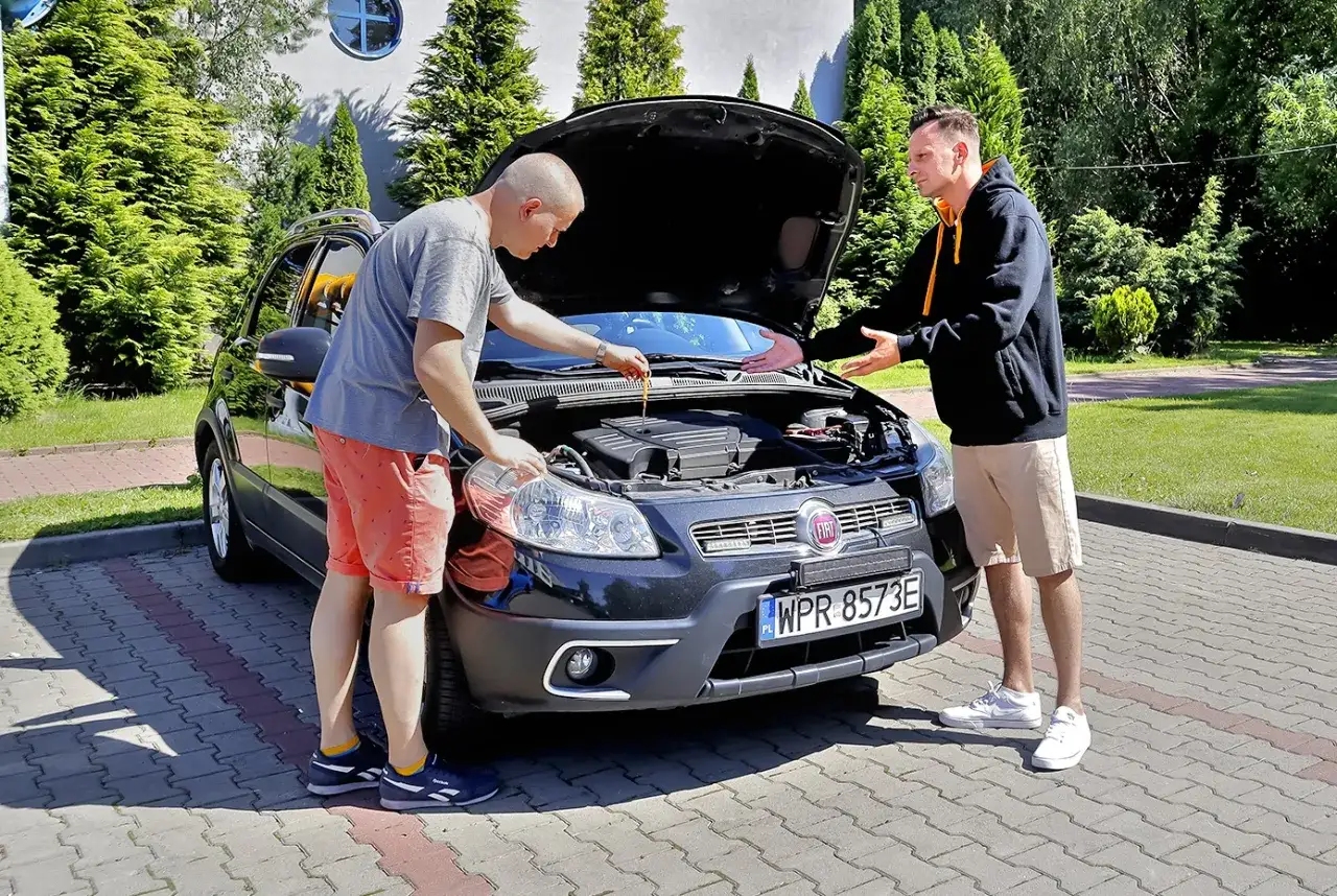 Dw&oacute;ch mężczyzn przy otwartej masce czarnego Fiata. Jeden sprawdza olej, drugi gestykuluje, jakby omawiał stan techniczny. To kluczowe przy negocjowaniu ceny auta.