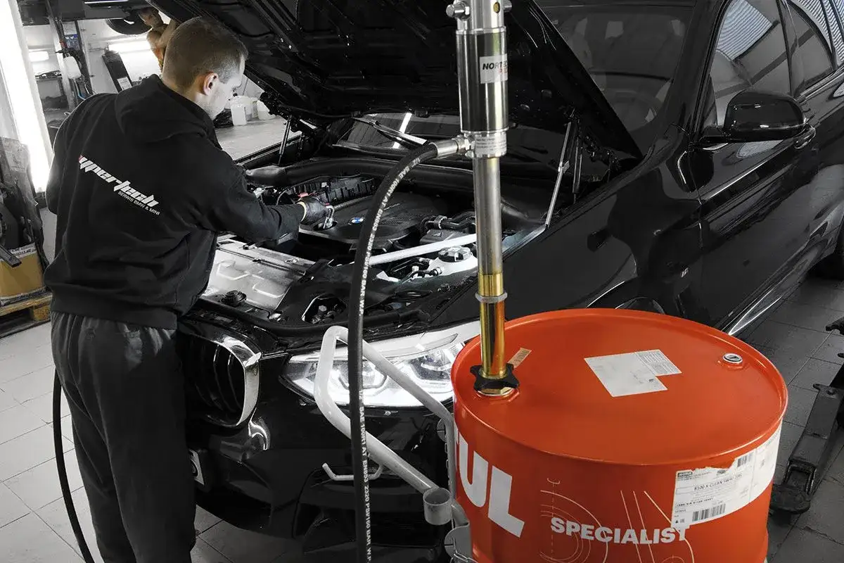 Mechanik wymienia olej w czarnym BMW. Obok stoi duży, pomarańczowy bęben z olejem. Regularna wymiana oleju co ile jest kluczowa dla silnika.