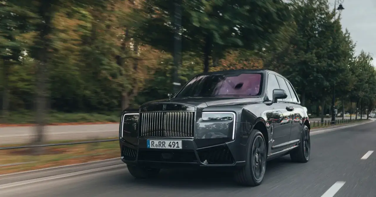 Czarny Rolls-Royce Cullinan, prawdopodobnie najdroższe auto w Polsce, pędzi po ulicy wśr&oacute;d drzew.