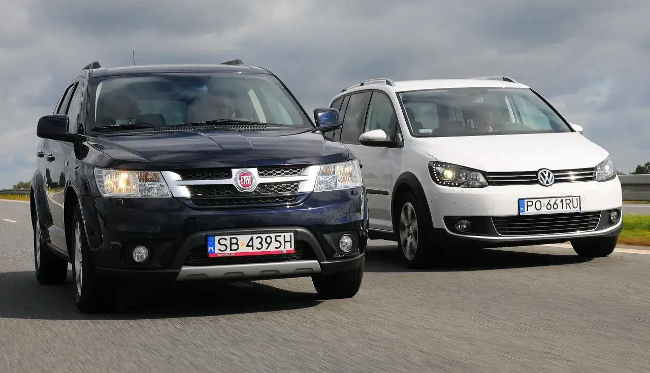 Ciemnoniebieski Fiat i biały Volkswagen, dwa duże samochody rodzinne, jadą obok siebie po drodze.