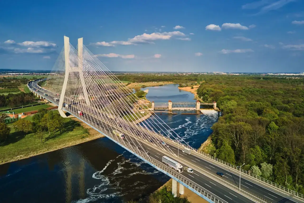 Widok z lotu ptaka na najdłuższy most w Polsce, imponującą konstrukcję z białymi pylonami i licznymi linami, przecinającą rzekę z widocznym w oddali mostem zwodzonym.