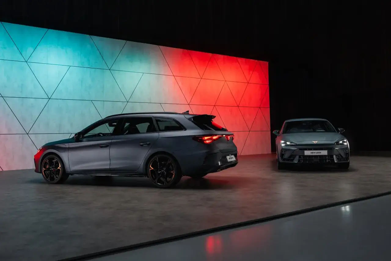 Dwa sportowe samochody Cupra Leon na tle ściany z geometrycznym wzorem, oświetlonej na niebiesko i czerwono. Cupra to marka dla tych, którzy szukają emocji.