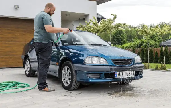 Mężczyzna myje niebieski samochód na posesji, używając węża ogrodowego. Woda spływa po karoserii i tworzy kałuże na podjeździe.