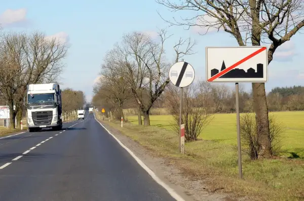 Znaki drogowe informują, ile można jechać poza terenem zabudowanym. Ciężarówka jedzie drogą, mijając pola i drzewa.