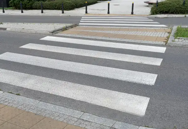 Sugerowane przejście dla pieszych z białymi pasami na szarym asfalcie, prowadzące przez chodnik i trawnik.