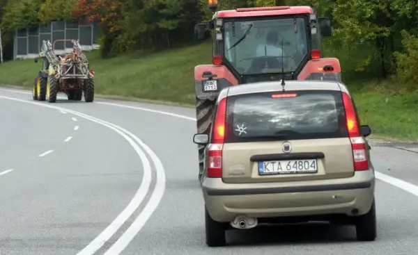 Złoty Fiat Panda jedzie za czerwonym traktorem. Czy można wyprzedzić traktor na podwójnej ciągłej? Droga kręta, drzewa po bokach.