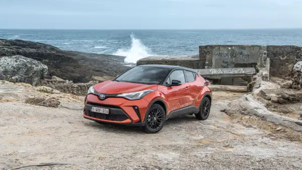 Pomarańczowe, małe auta hybrydowe Toyota C-HR na kamienistym wybrzeżu. W tle widać morze z falami i zniszczony budynek.