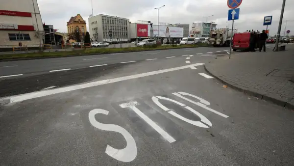 Na asfalcie widoczna jest biała linia warunkowego zatrzymania "STOP". W tle budynki i pojazdy.
