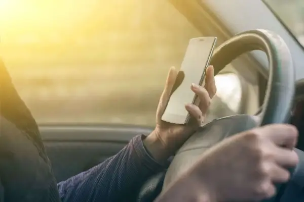 Ręka trzymająca telefon podczas jazdy samochodem. Słońce świeci przez szybę, oślepiając kierowcę.