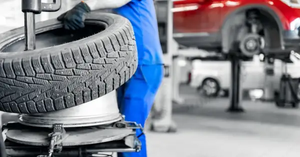 Mechanik w niebieskim kombinezonie trzyma oponę zimową. W tle widać podnośnik samochodowy i czerwony samochód. Sprawdź aktualne ceny wymiany opon.